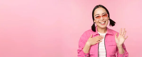 Retrato de chica adolescente asiática amigable en gafas de sol diciendo hola, agita su mano y sonríe, te saluda, gesto de hola, se para sobre fondo rosa —  Fotos de Stock