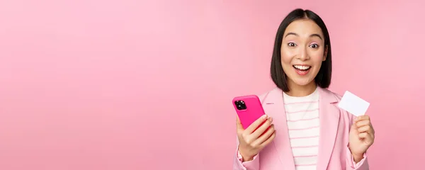 Imagen de la feliz mujer de negocios asiática sonriente mostrando tarjeta de crédito, pagando en línea en la aplicación del teléfono inteligente, orden con teléfono móvil, de pie sobre fondo rosa —  Fotos de Stock