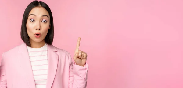 Retrato de mulher de negócios asiática, trabalhador corporativo de terno, parece surpreso com a propaganda, aponta dedo para cima, mostrando banner ou logotipo no topo, fica sobre fundo rosa — Fotografia de Stock