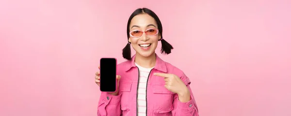 Mujer asiática entusiasta en ropa elegante, gafas de sol, apuntando con el dedo a la pantalla del teléfono móvil, mostrando la aplicación del teléfono inteligente, de pie sobre el fondo rosa —  Fotos de Stock
