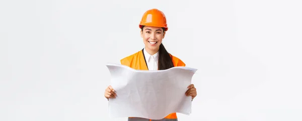 Lachend mooie Aziatische vrouwelijke architect, industriële vrouw in veiligheidshelm en reflecterende jas, tevreden na het lezen van blauwdrukken, het bouwen van huizen, iets bouwen, witte achtergrond — Stockfoto