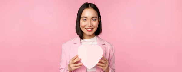 Questo è per te. Romantico carino asiatico donna d'affari, ragazza in abito, mostrando scatola a forma di cuore con regalo, in piedi su sfondo rosa — Foto Stock