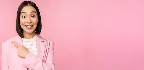 Retrato de mujer de negocios asiática, vendedora en traje apuntando con el dedo hacia la izquierda, mostrando el anuncio de la bandera, sonriendo y buscando profesional, fondo rosa —  Fotos de Stock
