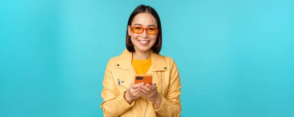 Smiling asian girl in sunglasses, using smartphone app, holding mobile phone, standing over blue background — Stock Photo, Image
