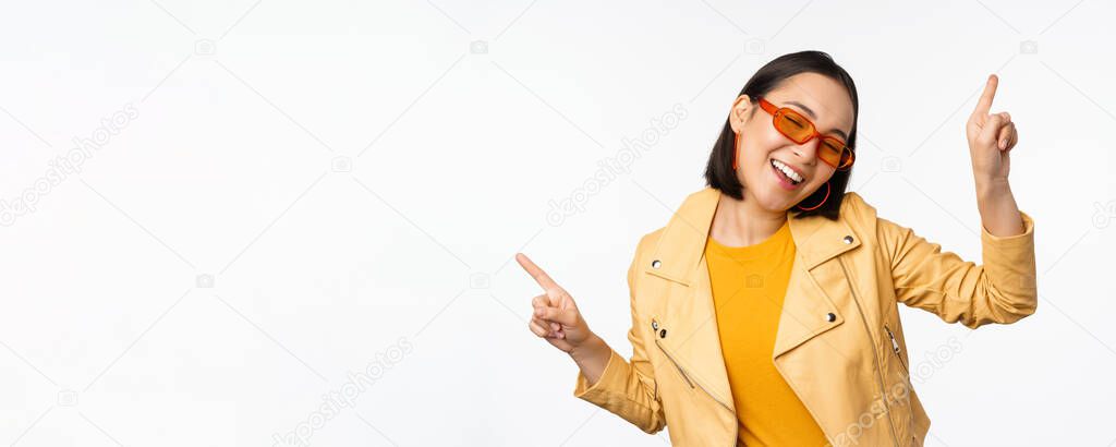 Smiling asian brunette woman in sunglasses, pointing fingers sideways, left and right, showing variants, laughing and dancing, wearing sunglasses, white background.