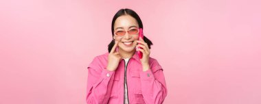 Güneş gözlüklü güzel Koreli kadın model, mutlu yüzlü cep telefonuyla konuşuyor, akıllı telefondan arkadaşını aramak için cep telefonunu kullanıyor, pembe arka planda duruyor.