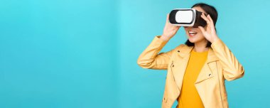 VR kulaklık takan mutlu Asyalı kadın modelin portresi. Sanal gerçeklik gözlükleriyle gülüyor ve gülümsüyor. Mavi arka planda duruyor.