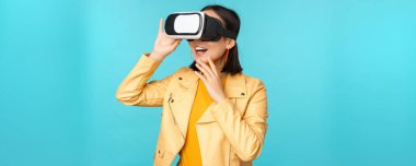 VR kulaklık takan mutlu Asyalı kadın modelin portresi. Sanal gerçeklik gözlükleriyle gülüyor ve gülümsüyor. Mavi arka planda duruyor.