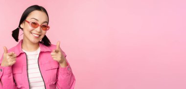 Güneş gözlüklü modern Asyalı kızın yakın portresi gülümsüyor, kamerayı işaret ediyor, seni övüyor, davet ediyor ya da iltifat ediyor, pembe arka planda duruyor.