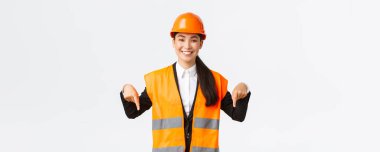 Gülümseyen neşeli inşaat müdürü, güvenlik kaskı takan Asyalı kadın mühendis ve yansıtıcı giysi anonsu. Kadın proje yöneticisi aşağı işaret ediyor, beyaz arka planda duruyor