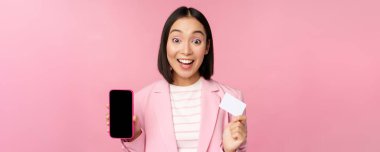 Ateşli Asyalı iş kadını cep telefonu ekranı ve kredi kartı gösteriyor, kameraya hayran bakıyor, pembe arka planda duruyor.