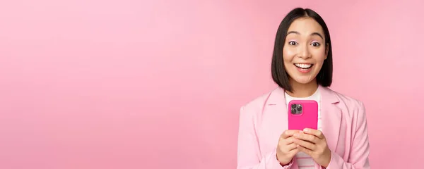 Porträtt av asiatisk flicka i kostym med smartphone, leende och ser glad, står över rosa studio bakgrund — Stockfoto