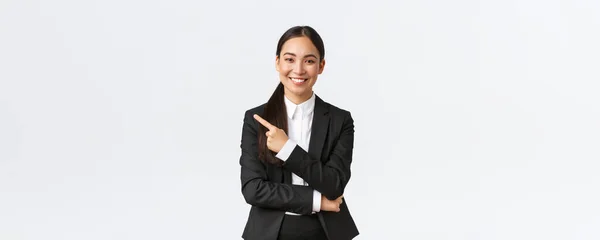 Щаслива професійна азіатська жінка-менеджерка, бізнес-леді в костюмі, що показує оголошення, посміхнений і вказує палець, що залишився за продуктом або банером проекту, стоїть на білому тлі — стокове фото