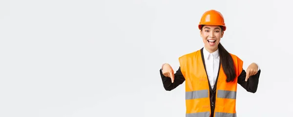 Izgalmas mosolygós ázsiai mérnök, ipari nő biztonsági sisakban és fényvisszaverő kabátban, ujjal mutogatva lefelé, bejelentve. Építész bemutató neki projekt, fehér háttér — Stock Fotó