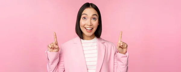 Захоплений корпоративний працівник, азіатська бізнес-леді спрямовує пальці вгору і посміхається, показуючи рекламу, логотип, стоїть на рожевому фоні — стокове фото