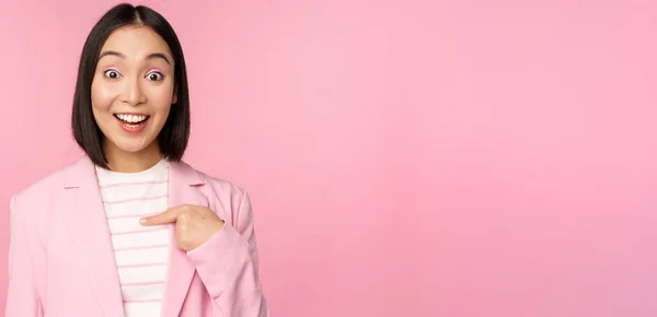 Портрет молодої азіатської бізнес-леді з з здивованим, збудженим виразом обличчя, вказуючи пальцем на себе, стоячи в костюмі над рожевим тлом — стокове фото