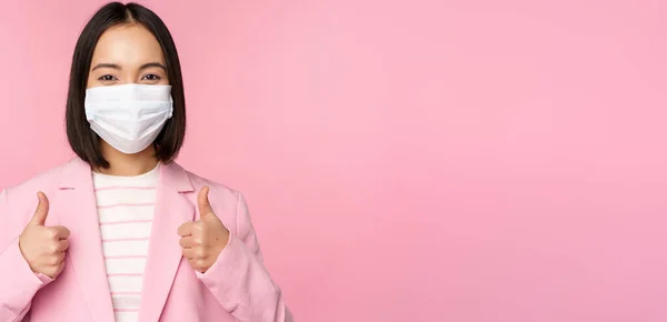 Empresária asiática de terno e máscara facial médica, mostrando os polegares para cima, recomendando o uso de equipamentos de proteção individual no escritório durante a pandemia covid-19, fundo rosa — Fotografia de Stock