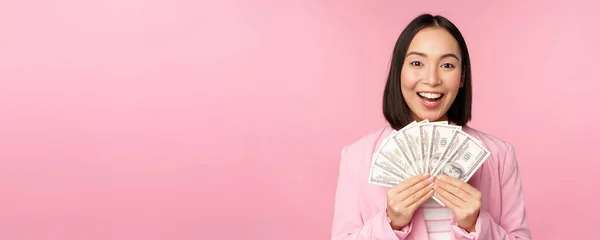 Ekonomi, mikrokrediter och människor koncept. Glad leende asiatisk affärskvinna visar dollar pengar, står i kostym mot rosa bakgrund — Stockfoto