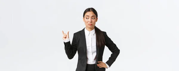 Scettico e imbarazzante giovane donna d'affari asiatica, commessa in abito nero modellare qualcosa di piccolo e cercando deluso in termini di dimensioni, smorzare indivertito su piccola cosa, sfondo bianco — Foto Stock