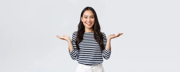 Estilo de vida, emociones de la gente y concepto casual. Linda mujer asiática sonriente introducir dos productos, tomarse de las manos de lado como si demostrara productos en las palmas de las manos, de pie fondo blanco —  Fotos de Stock