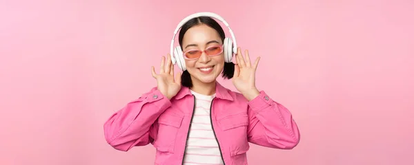 Танцююча стильна азіатська дівчина слухає музику в навушниках, позує на рожевому фоні — стокове фото