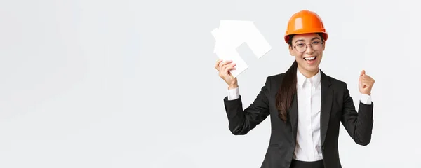 Successful winning asian female construction engineer, team lead architect achieve goal, holding house maket and fist pump in rejoice, celebrating victory in helmet and business suit — Stock Photo, Image