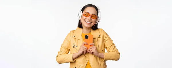 Concept technologique. Élégante fille asiatique dans les écouteurs, tenant smartphone, danse et chant, écouter de la musique, debout sur fond blanc — Photo