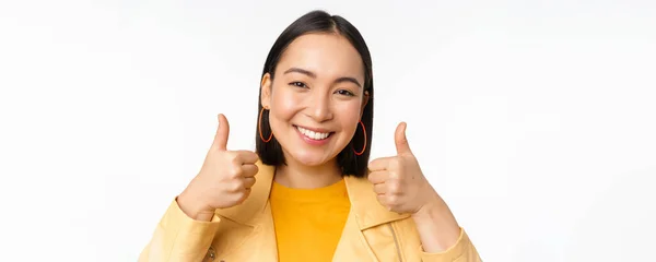 Smiling asian girl showing thumbs up, looking pleased, approve smth, standing over white background — Stock Photo, Image