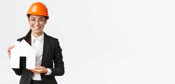 Lächelnd gut aussehende asiatische Verkäuferin einer Baufirma, die Eigenheime verkauft, trägt Helm und Businessanzug, zeigt ein gemütliches Zuhause, stehender weißer Hintergrund — Stockfoto