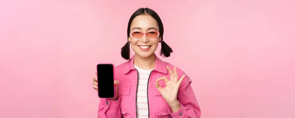 Entusiasta joven mujer asiática mostrando bien, ok signo, sonriendo complacido, pantalla del teléfono móvil, aplicación de teléfono inteligente, de pie sobre fondo rosa —  Fotos de Stock