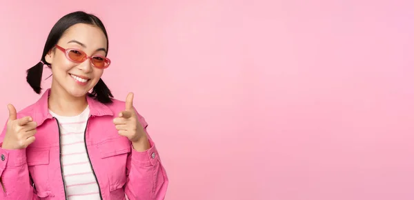 Крупним планом портрет сучасної азіатки в сонцезахисних окулярах посміхається, вказуючи пальці на камеру, хвалити вас, запрошуючи або компліментуючи, стоячи над рожевим тлом — стокове фото