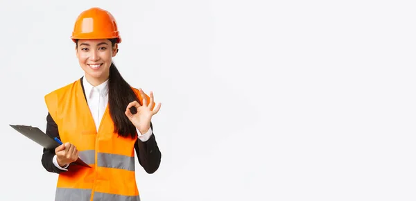 Tevreden glimlachende Aziatische vrouwelijke architect, bouwkundig ingenieur in helm en reflecterende jas, tonen oke gebaar, goedkeuren en toestemming geven voor het bouwen van werken, afwerking inspectie — Stockfoto