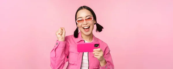 Feliz chica coreana sonriente ganando en el teléfono móvil, mirando la pantalla horizontal del teléfono inteligente y regocijándose, lograr la meta, celebrando, de pie sobre el fondo rosa — Foto de Stock