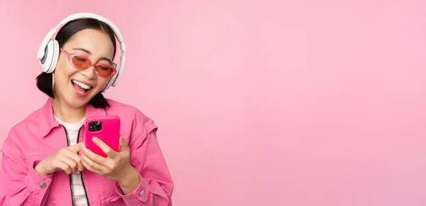 Stylowa azjatycka dziewczyna tańcząca ze smartfonem, słuchająca muzyki w słuchawkach w aplikacji na telefon komórkowy, uśmiechnięta i śmiejąca się, pozująca na różowym tle — Zdjęcie stockowe