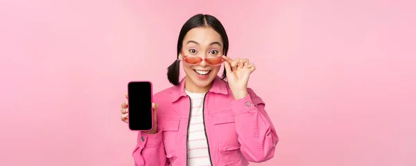 Портрет азійської дівчини, що показує екран мобільного телефону, реагує здивовано, стоячи над рожевим фоном. — стокове фото