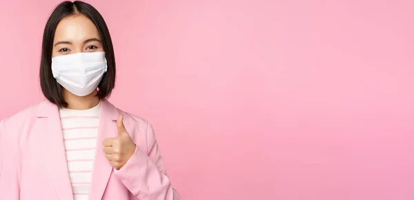 Asiatisk affärskvinna i kostym och medicinsk ansiktsmask, visar tummen upp, rekommenderar bär personlig skyddsutrustning på kontoret under covid-19 pandemi, rosa bakgrund — Stockfoto