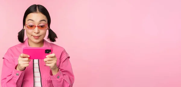 Porträtt av glad asiatisk flicka som spelar på smartphone, titta på videor på mobiltelefon app, står över rosa bakgrund — Stockfoto