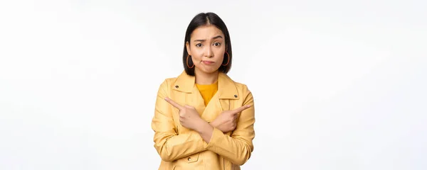 Afbeelding van besluiteloos aziatisch meisje, wijzend vingers zijwaarts, wijzend naar links en rechts, kiezen variant, beslissen, staande over witte achtergrond — Stockfoto