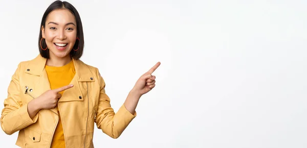 Entusiastiska asiatiska flicka pekar finger höger, visar reklam och leende, inbjudande till butiken, står över vit bakgrund — Stockfoto