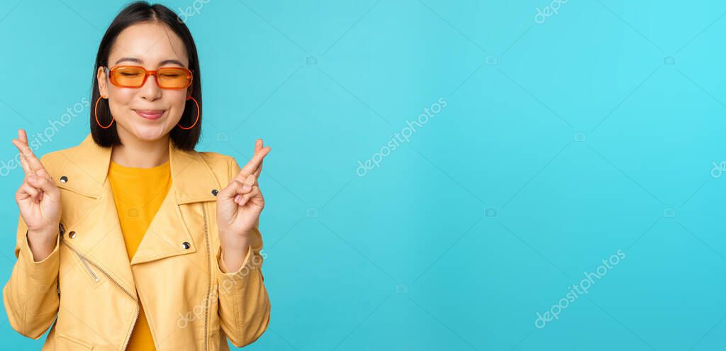 Smiling beautiful asian woman wishing, cross fingers for good luck and looking hopeful, standing over blue background. Copy space