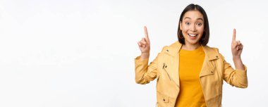 Hevesli Asyalı kız parmaklarını havaya kaldırıyor, üstte reklamını gösteriyor, mutlu bir şekilde gülümsüyor, tanıtım teklifi ya da afiş sunuyor, beyaz arka planda duruyor.