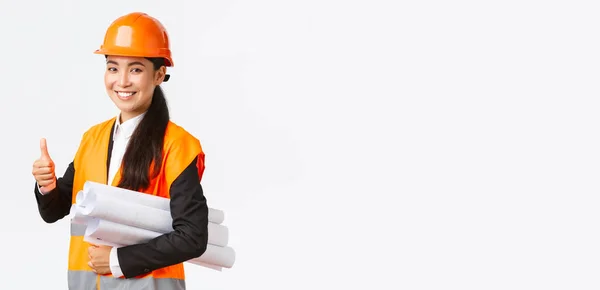 Σίγουρη επιτυχημένη γυναίκα ασιατική διευθυντής κατασκευών, αρχιτέκτονας σε κράνος και σακάκι ασφαλείας, δείχνει αντίχειρα-up και μεταφέρουν τα σχέδια του έργου του κτιρίου, εγγυώνται την ποιότητα, λευκό φόντο — Φωτογραφία Αρχείου