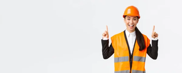 Edilizia, costruzione e concetto industriale. Felice sorridente ingegnere asiatico femminile in casco di sicurezza e abbigliamento riflettente, introdurre nuovo oggetto, tenuta in vendita. Architetto che punta il dito verso l'alto — Foto Stock