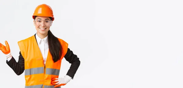 Sonriente alegre asiático mujer construcción manager introducir su interprise para los inversores, usando gafas de seguridad, casco y guantes apuntando mano izquierda y buscando orgulloso, fondo blanco — Foto de Stock