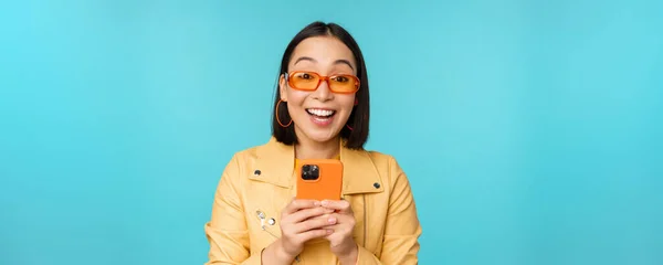 Porträtt av entusiastisk asiatisk kvinna i solglasögon, med mobiltelefon, ler och skrattar, ser glad, håller smartphone, står över blå bakgrund — Stockfoto