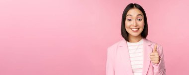 Asyalı iş kadını portresi tatmin olmuş gülümsüyor, başparmaklarını gösteriyor, övüyor ve onaylıyor, pembe arka planda takım elbise içinde duruyor.