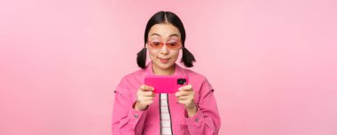 Akıllı telefondan oynayan mutlu Asyalı kızın portresi, cep telefonu uygulamasında videolar izlemek, pembe arka planda durmak.