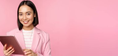Asyalı genç kadın portresi, dijital tabletli ofis hanımı, takım elbiseli, gülümseyen ve profesyonel görünen, pembe arka planda poz veren