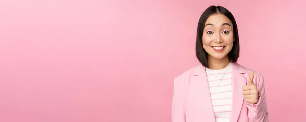 Portrait de femme d'affaires asiatique souriant satisfait, montrant pouces levés, louanges, comme et approuver, debout en costume sur fond rose — Photo