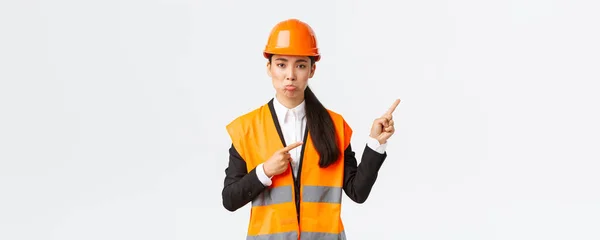 Construção, construção e conceito industrial. Desapontado e desapontado mulher asiática gerente, engenheiro em segurança capacete puff, reclamando algo ruim, apontando dedos canto superior direito — Fotografia de Stock
